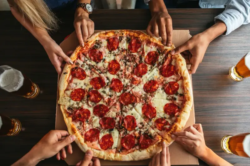 Hände greifen nach einer großen Salamipizza, die auf einem Tisch liegt. Jeder bekommt seinen fairen Anteil an pizzateig pro person.