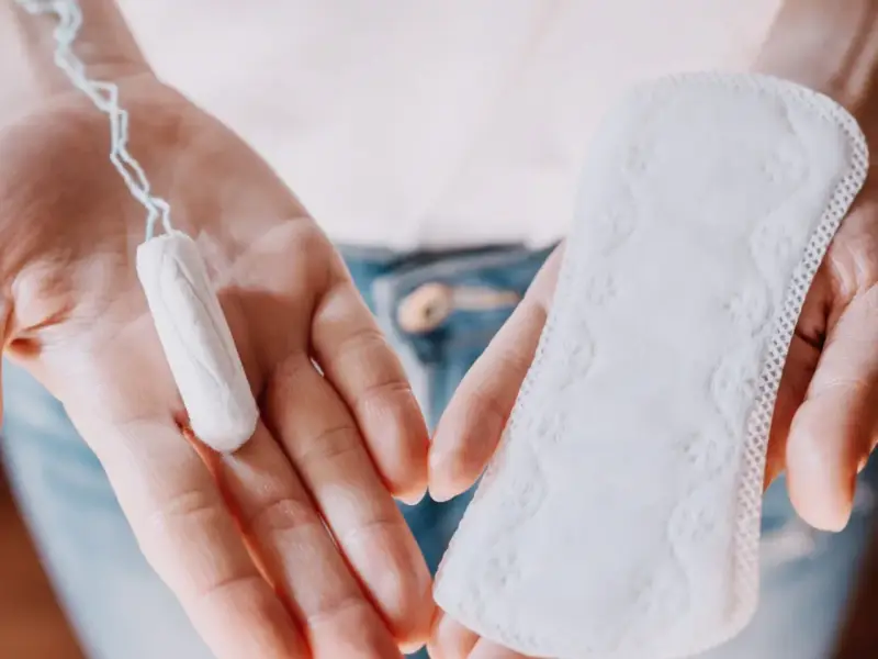 Dłonie trzymają tampon i podpaskę. Czasem pojawia się plamienie zamiast okresu, wtedy wybór jest ważny.