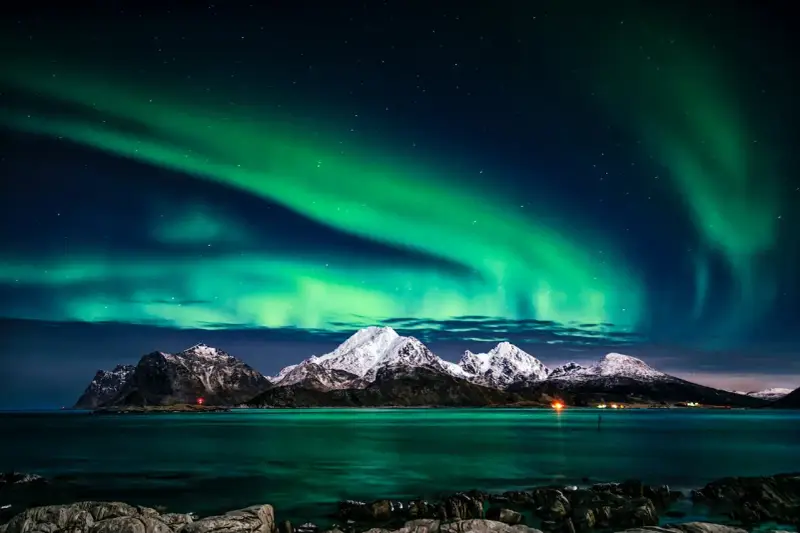Zielona zorza polarna nad ośnieżonymi górami i morzem. Marzysz, jak dojechać do Norwegii, by zobaczyć to na żywo?