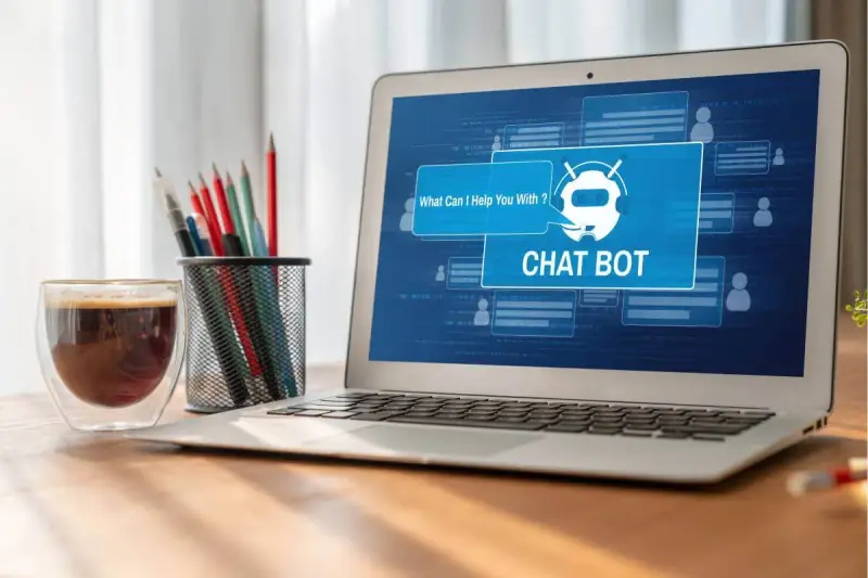 Na ekranie laptopa widać interfejs chatbota, który oferuje pomoc. Obok stoi szklanka kawy i pojemnik z przyborami do pisania.