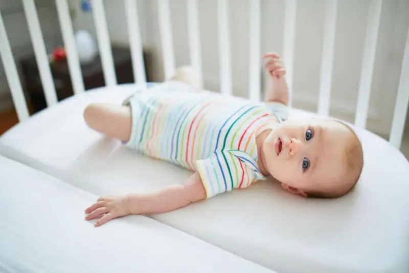 Niemowlę w kolorowym body leży w łóżeczku. Jego niebieskie oczy przypominają **perły Epsteina**, a uśmiech rozjaśnia pokój.