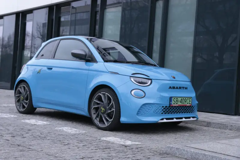 Niebieski Abarth 500e, gotowy do testów, zaparkowany przed nowoczesnym budynkiem.