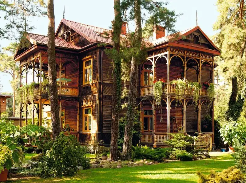 Drewniana willa w stylu świdermajer, otoczona zielenią, z pięknymi werandami i zdobieniami.