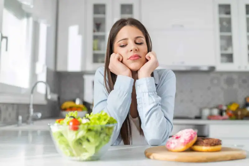 Femme triste face à une salade et des donuts, pensant aux aliments à éviter pour la dermite séborrhéique.