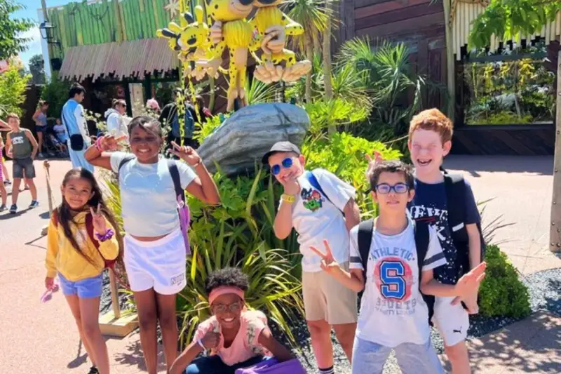Des enfants joyeux font des signes de paix devant une attraction, un avis "supernova juniors" est visible.