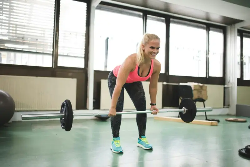En kvinna lyfter skivstång i ett gym. Hon ler och ser fokuserad ut, en bra start på hur blir man stark.