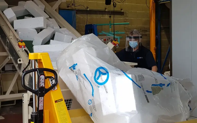 Pracownik w masce i przyłbicy przygotowuje styropian do recyklingu. Duży worek z logo Knauf Circular symbolizuje proces recyklingu styropianu.