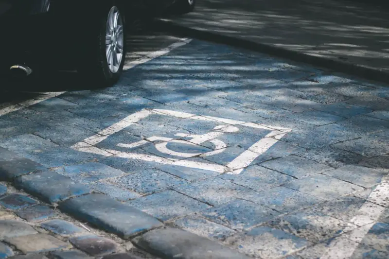 Niebieskie miejsce parkingowe dla niepełnosprawnych pod blokiem, z symbolem wózka inwalidzkiego. Obok widoczna część czarnego samochodu.