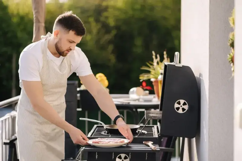 Mężczyzna w fartuchu przygotowuje mięso na grillu na balkonie. Czy można robić grilla na balkonie? Tak, jeśli jest bezpiecznie!