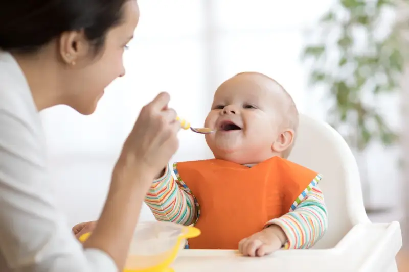 Mama karmi uśmiechniętego niemowlaka łyżeczką. To idealny przykład, co na śniadanie dla niemowlaka – zdrowo i radośnie!