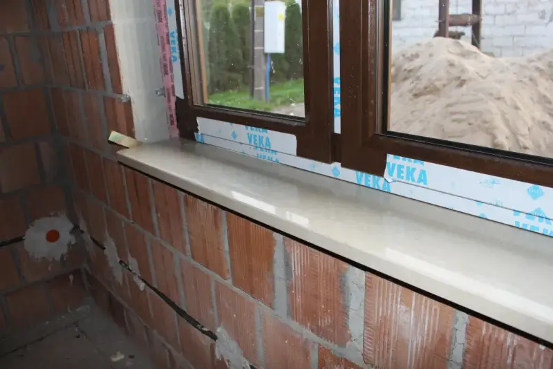 Montaż parapetów zewnętrznych po ociepleniu. Nowy, jasny parapet lśni pod brązową ramą okna, a ceglana ściana czeka na dalsze prace.