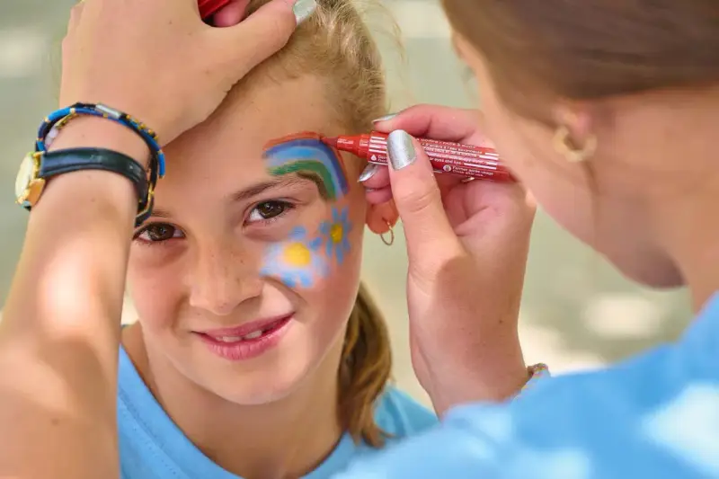 Une petite fille souriante se fait maquiller sur la joue un arc-en-ciel et des fleurs, au stand maquillage d'une kermesse.