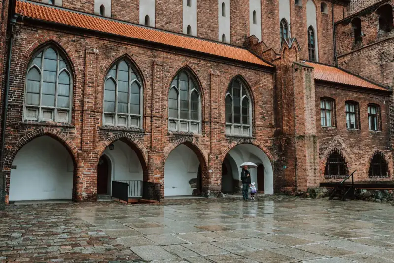 Zamek w Kwidzynie, z ceglanymi arkadami i gotyckimi oknami, skrywa tajemnice. Dwoje ludzi z parasolem spaceruje po mokrym dziedzińcu.