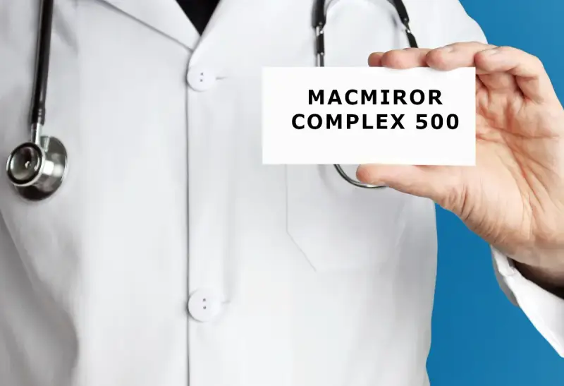 Lekarz w białym kitlu trzyma kartkę z napisem MACMIROR COMPLEX 500. Stetoskop wisi na jego szyi.
