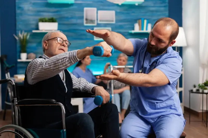Starszy mężczyzna na wózku wykonuje ćwiczenia z hantlami pod okiem fizjoterapeuty. To kluczowy element rehabilitacji po udarze.