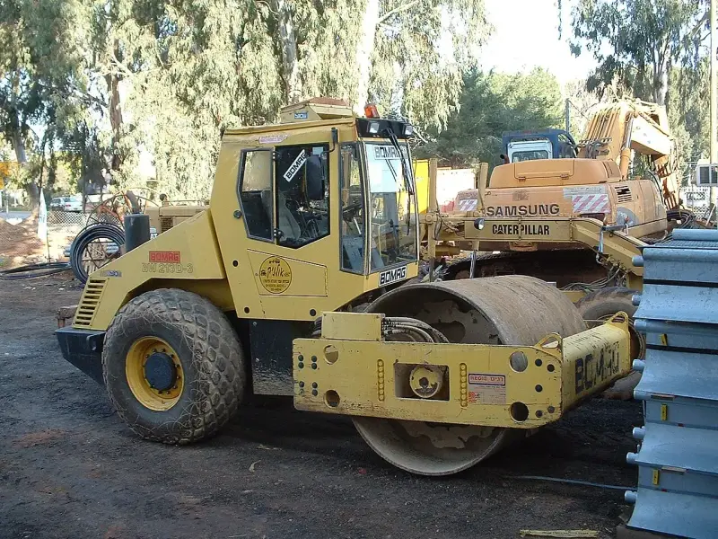 Żółty walec drogowy BOMAG BW 213D-3 gotowy do budowy drogi. W tle koparka Samsung Caterpillar.