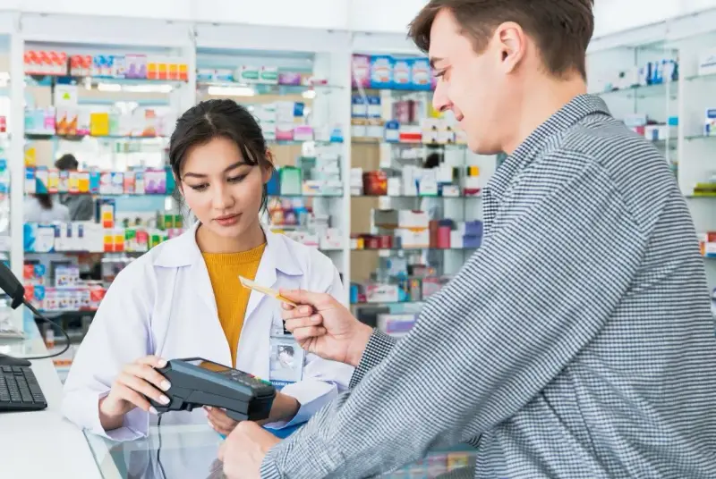 Klient płaci kartą w aptece, gdzie farmaceutka obsługuje terminal. W tle widać półki z lekami, a na nich produkt z napisem ursopol.