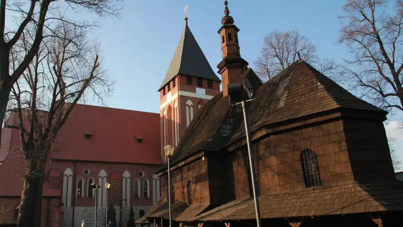 Drewniana i ceglana architektura kościołów w miasteczku śląskim, z wieżą zegarową i strzelistymi dachami, pod błękitnym niebem.