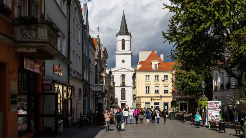 Ulica w jednym z miast na zachodzie Polski, z kościołem w tle i ludźmi spacerującymi po rynku.