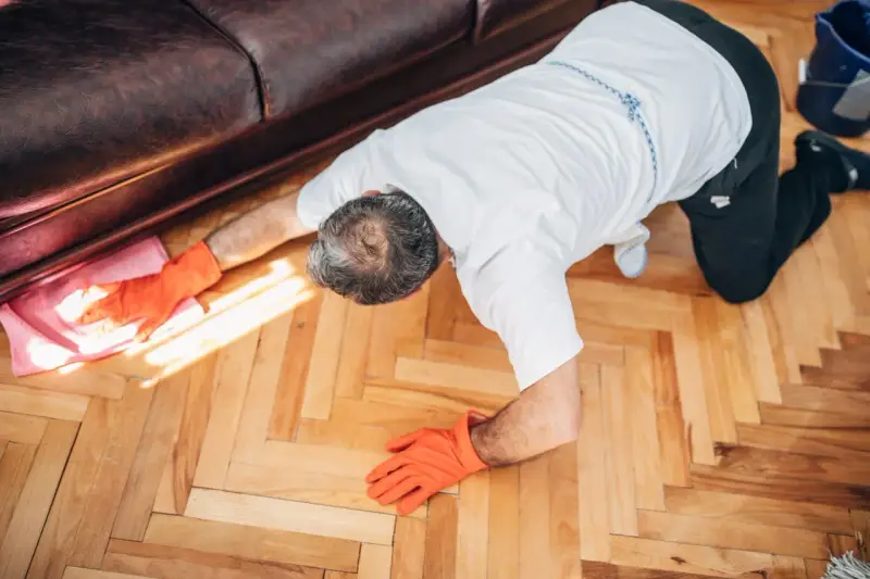 Man in orange gloves cleaning wooden floor with a pink cloth.