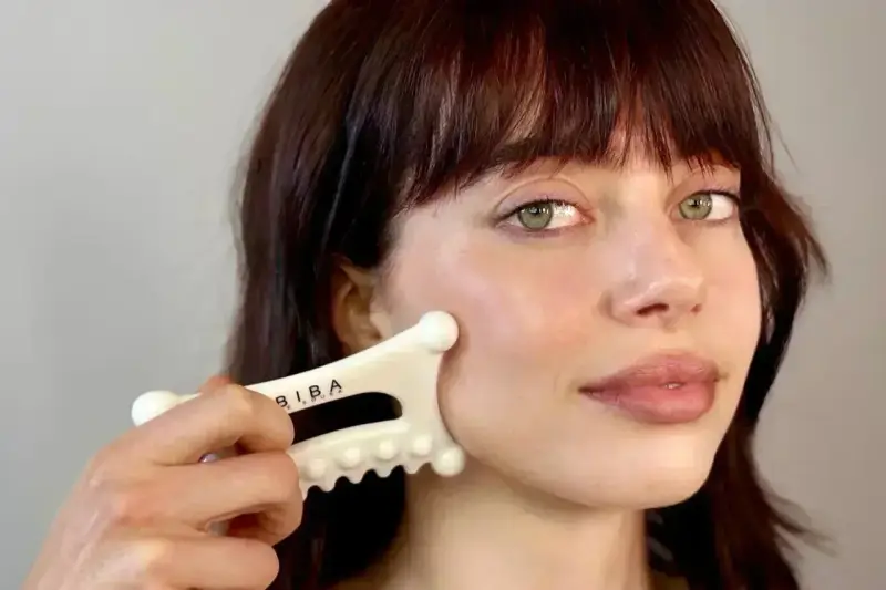 A woman with bangs and green eyes performs a facial fascia massage using a white gua sha tool.