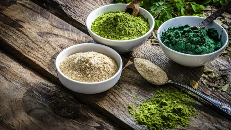 Drei Schalen mit Pulver auf Holz: hellbraun, leuchtend grün und dunkelgrün. Spirulina gesund und voller Nährstoffe.