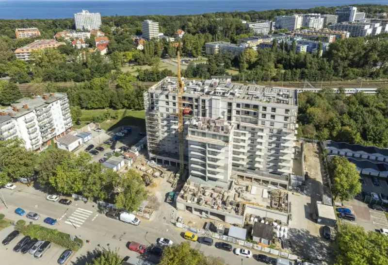 Budowa bloku mieszkalnego w Kołobrzegu. Widok z lotu ptaka na plac budowy, dźwig, samochody i okoliczne budynki. Ceny mieszkań Kołobrzeg mogą być różne.