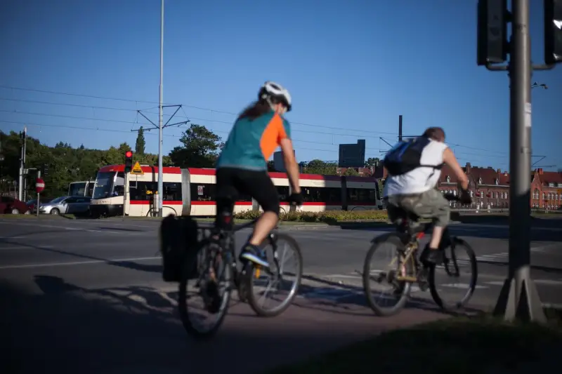 Rowerzyści jadący wzdłuż torów tramwajowych, w tle czerwono-biały tramwaj. Trasa rowerowa wiodąca w kierunku Westerplatte.