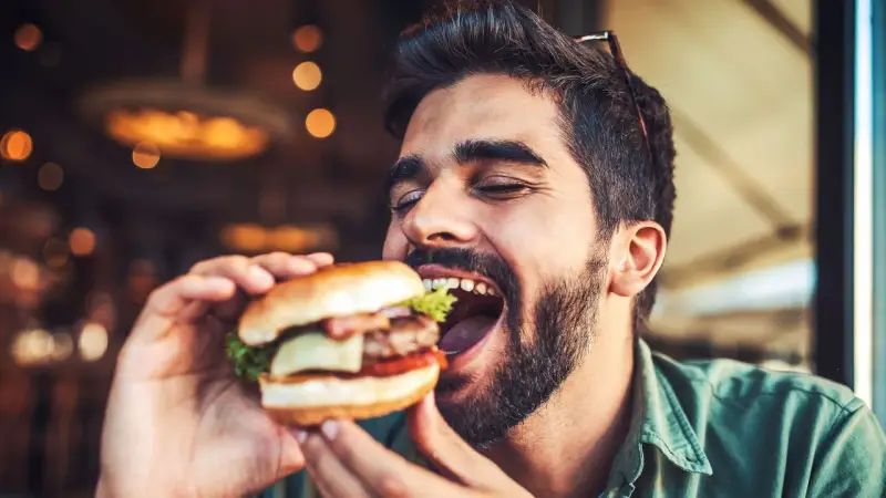 Hombre disfrutando un jugoso **cheat meal** de hamburguesa, con los ojos cerrados por el placer.