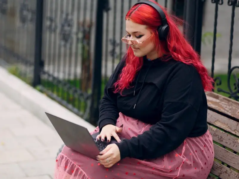 Młoda babista z ognistymi włosami i słuchawkami na uszach pracuje na laptopie, siedząc na ławce w parku.