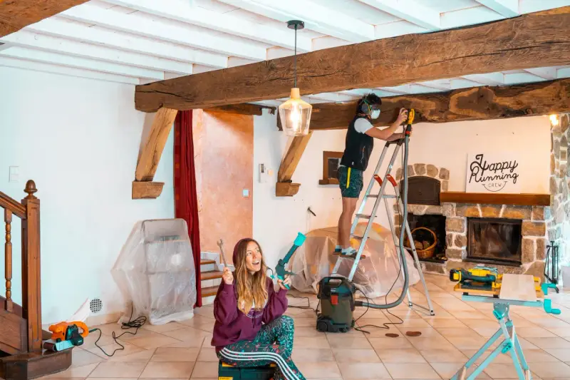 Un homme ponce une poutre en bois avec une ponceuse, tandis qu'une femme tient des outils.