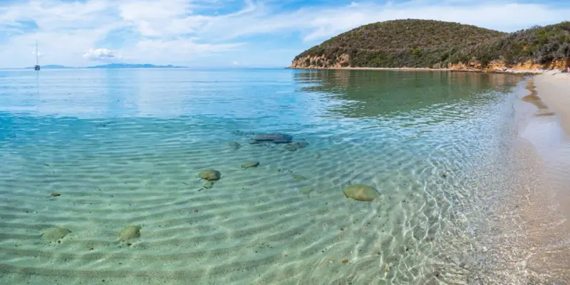 Piękne livorno plaże z krystalicznie czystą wodą i piaszczystym dnem. W oddali widać żaglówkę i zielone wzgórza.