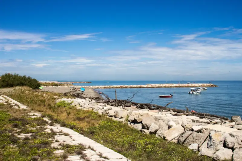 Sentiero in pietra e mare calmo con barche. Per capire come arrivare a Pellestrina da Fusina, immagina di percorrere questa costa.