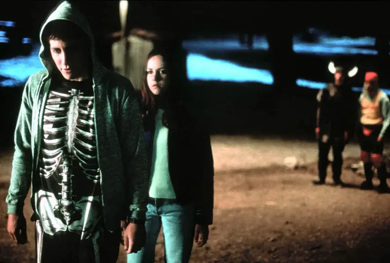 Donnie Darko w bluzie z kapturem i koszulce z nadrukiem szkieletu, obok dziewczyny. W tle dwie postacie w kostiumach.
