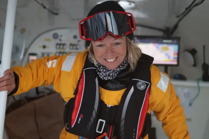 Alexia Barrier, souriante, porte une combinaison jaune et un gilet de sauvetage, prête pour une aventure maritime.