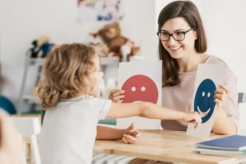 Dziecko i pedagog pracują nad emocjami podczas terapii pedagogicznej, używając kart z buźkami.