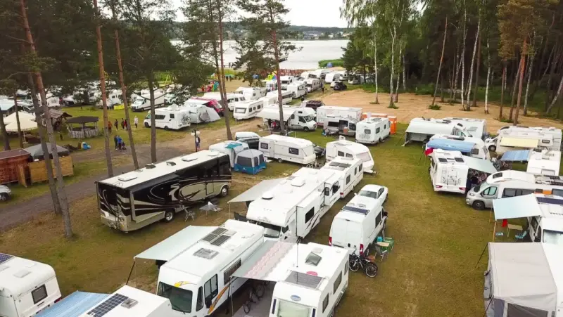 Duży zlot caravaningowy nad jeziorem. Wiele kamperów i przyczep kempingowych zaparkowanych na trawie, otoczonych drzewami.