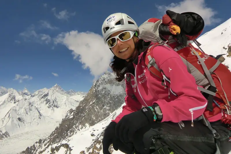 Gerlinde Kaltenbrunner, lächelnd in voller Bergsteigerausrüstung, vor einer majestätischen Schneelandschaft.