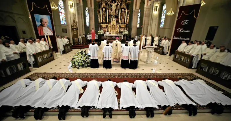 Klerycy w białych szatach leżą na posadzce kościoła, w geście pokory. Obok nich stoją inni duchowni, a w tle widać ołtarz i portret papieża.