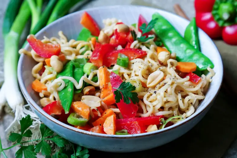 Bunter mie nudelsalat mit Erdnüssen, Zuckerschoten, Karotten und Paprika, garniert mit Petersilie.
