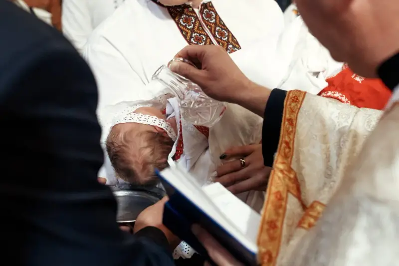 Ceremonia chrztu dziecka. Duchowny polewa główkę niemowlęcia wodą święconą. Warto wiedzieć, ile kosztuje chrzest.