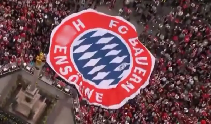 Ogromna flaga z herbem FC Bayern Monachium niesiona przez tłum kibiców.