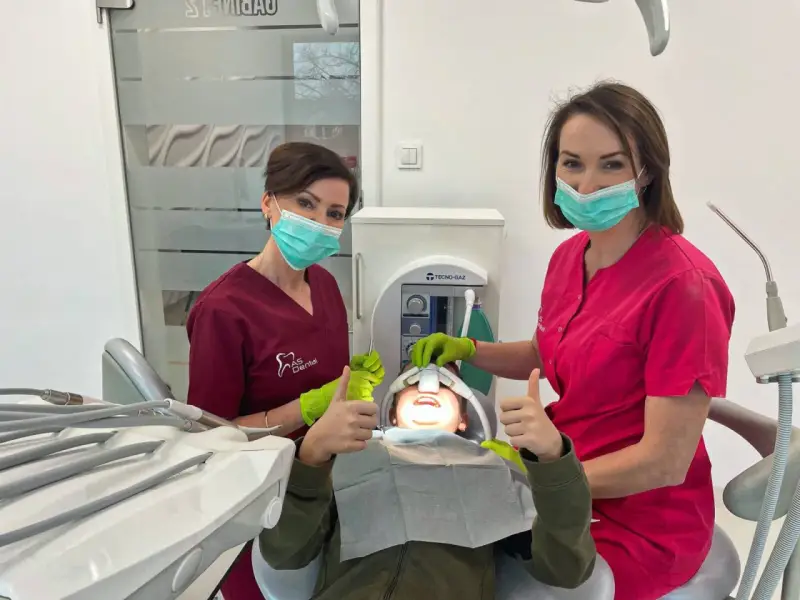 Dwie dentystki w maseczkach i rękawiczkach z uśmiechniętym pacjentem w gabinecie stomatologicznym.