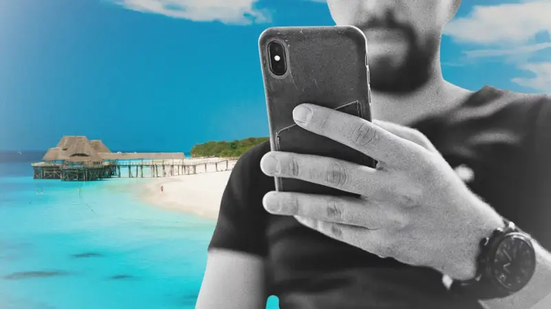 Mężczyzna z telefonem na plaży na Zanzibarze, sprawdzający zanzibar internet. W tle turkusowa woda i drewniany pomost.