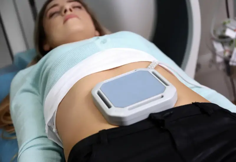 Kobieta leży podczas zabiegu, na brzuchu ma nałożone urządzenie do terapii polem magnetycznym, co jest częścią jej rehabilitacji.
