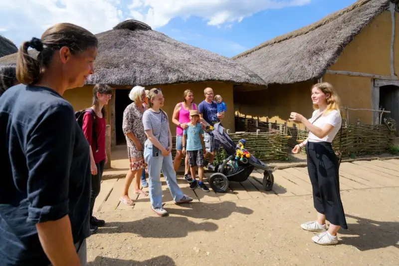 Familienurlaub in Haithabu: Eine Gruppe erkundet historische Häuser mit Strohdächern, während eine junge Frau eine Führung gibt.