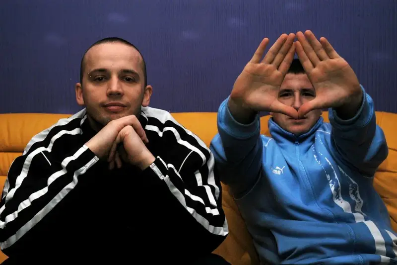 Dwóch mężczyzn w dresach, jeden z rękami złożonymi na brodzie, drugi tworzy rękami kształt serca. Klimat hip hop i rap.