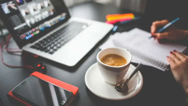Tazza di caffè, laptop, smartphone e mani che scrivono su un quaderno. Questo è cosa significa da remoto: lavorare ovunque.