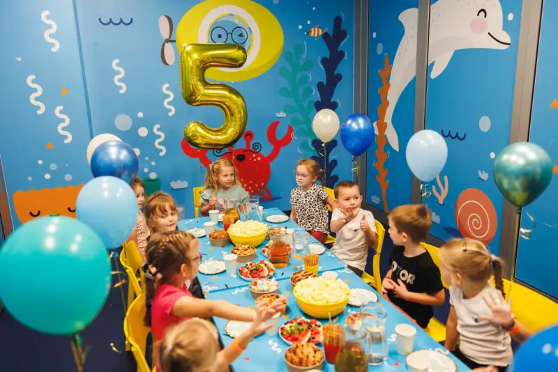 Dzieci świętują 5 urodziny przy stole pełnym przekąsek. Złoty balon w kształcie cyfry 5 unosi się nad stołem.