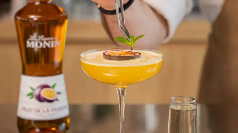 Barman dekoruje drink likierem z marakui, dodając listek mięty. Obok stoi butelka Monin.